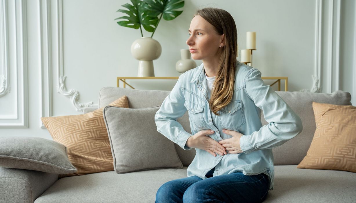 Junge Frau sitzt auf dem Sofa und hält sich den Bauch.