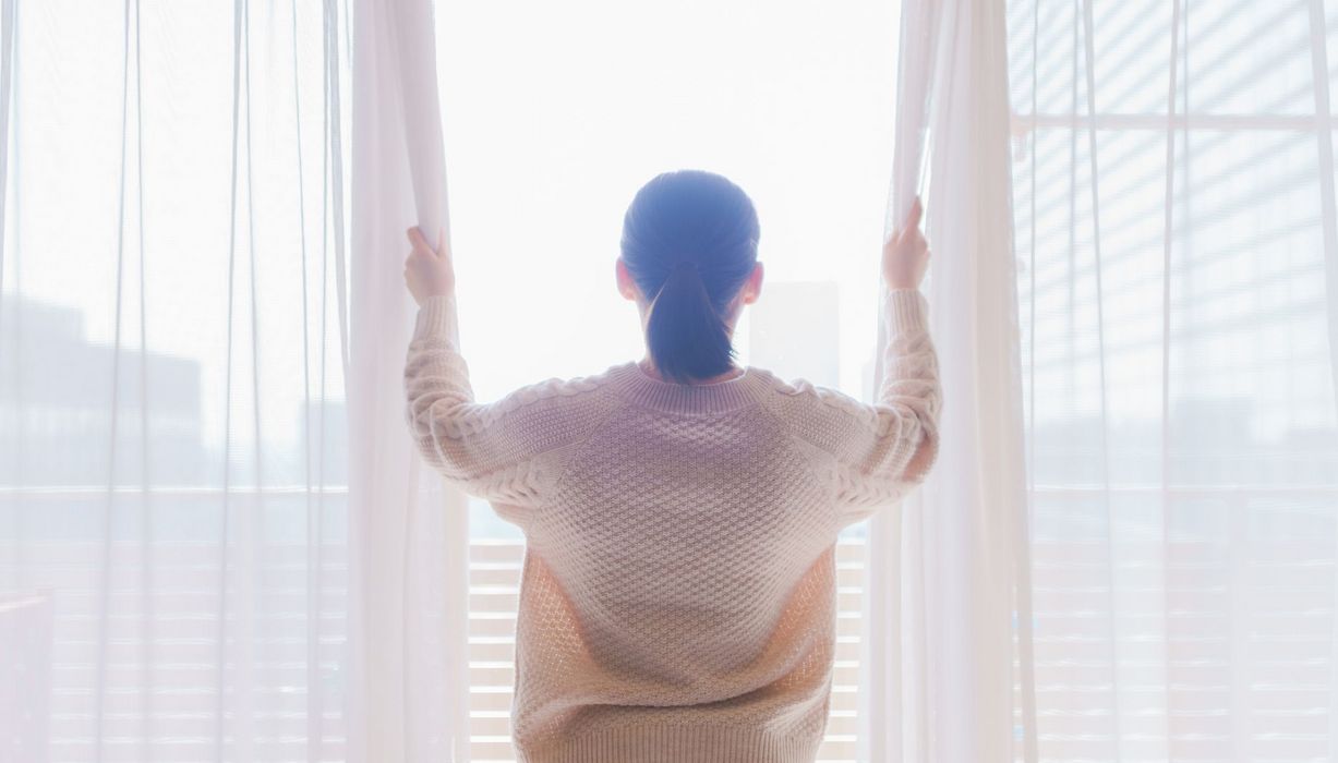 Frau steht mit dickem Wollpullover am Fenster und öffnet die Vorhänge.