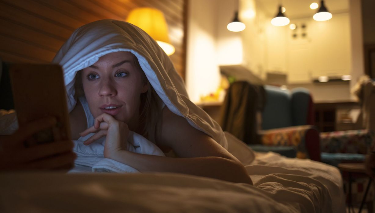 Junge Frau, liegt im Bett und schaut auf ihr Smartphone/Tablet.