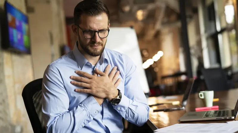 Junger Mann, sitzt im Büro und hält seine beiden Hände an sein Herz.