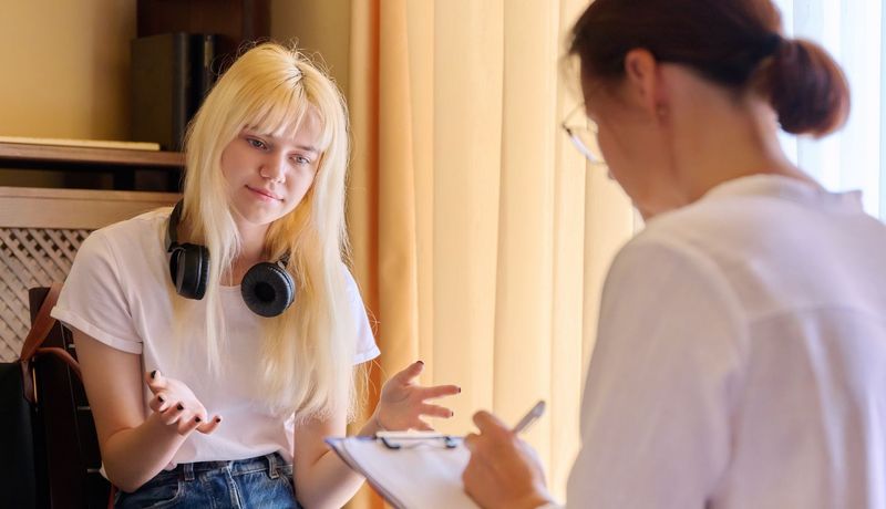 Jugendliche mit Kopfhörern um den Nacken gestikuliert. Ihr gegenüber sitzt eine Frau, die sich Notizen auf einem Klemmbrett macht.