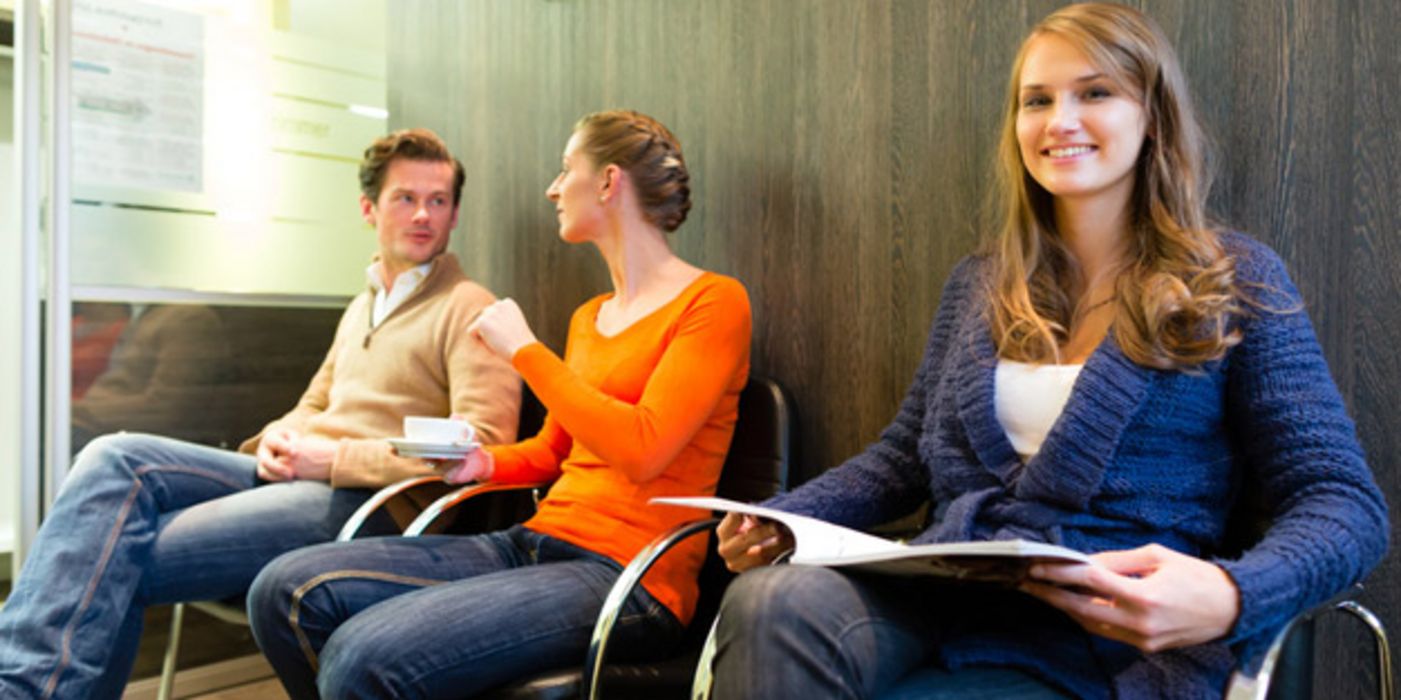 Zwei junge Frauen und ein junger Mann sitzen im Wartezimmer einer Arztpraxis, einer der Frauen schaut in die Kamera