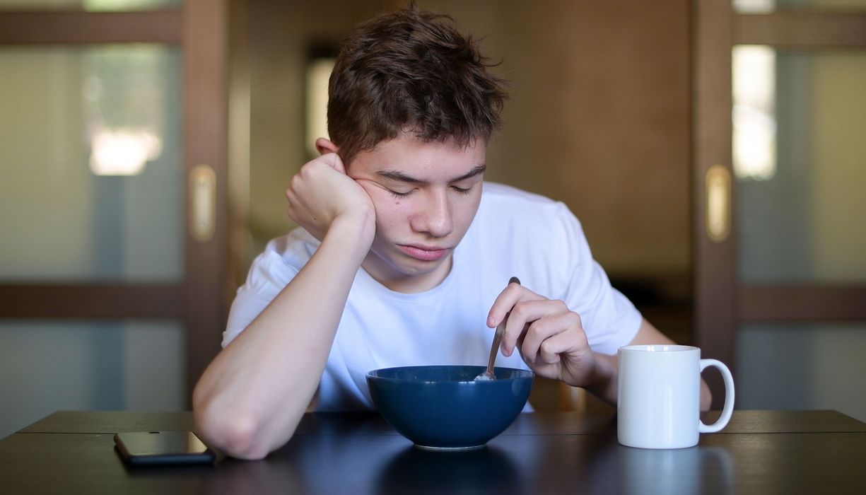 Jugendlicher, sitzt morgens sehr müde vor seinem Müsli und stochert darin herum.