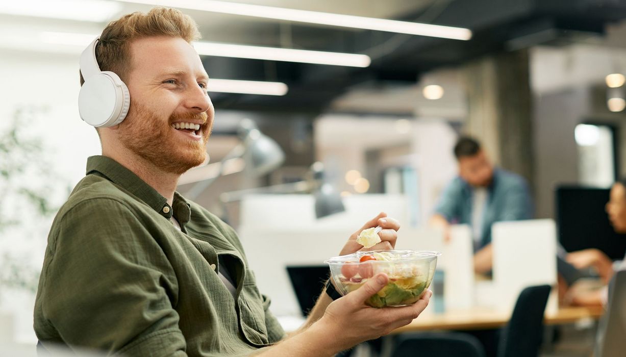 Junger Mann mit Kopfhörer isst Müsli im Büro.