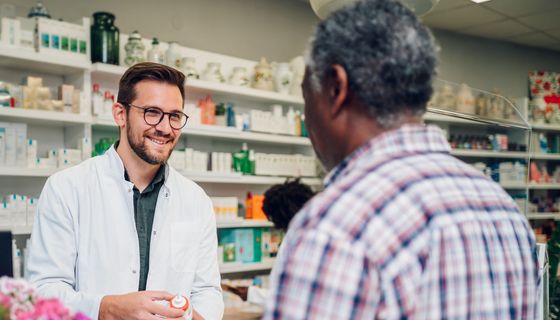 Ein lächelnder Apotheker reicht einem Kunden eine Arzneimittel. Im Hintergrund sind Regale mit weiteren Medikamenten zu sehen.