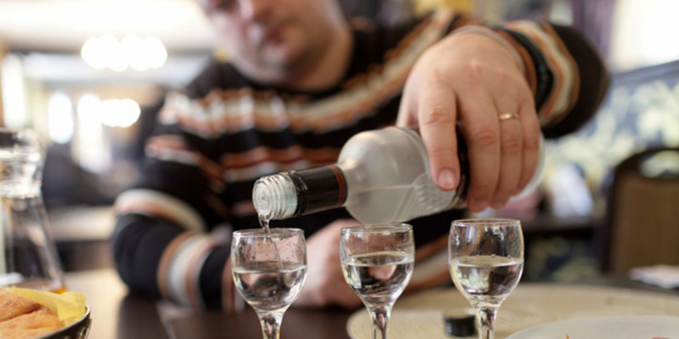 Gedeckter Tisch, im Vordergrund Vodkagläser,die von einem im unscharfen Hintergrund sitzenden Mann vollgeschenkt werden.