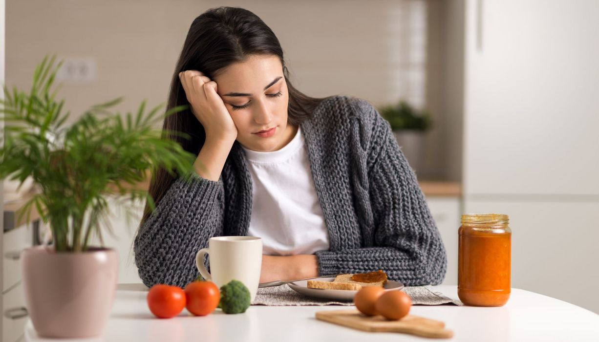 Junge Frau, sitzt vor einem Tisch mit Brot, Eiern und Gemüse und schaut verzweifelt nach unten.