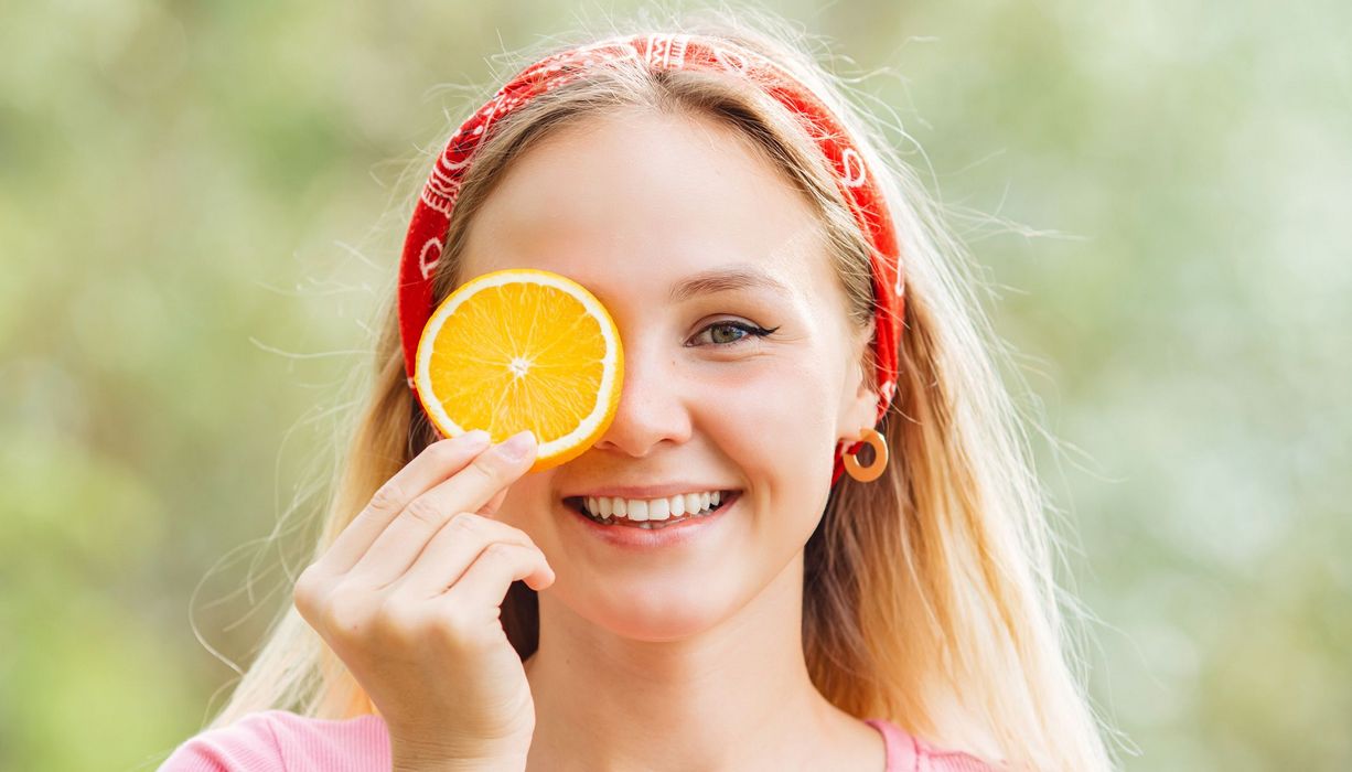 Junge Frau, hält sich eine Orangenscheibe vor das Auge und lacht.