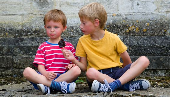 Zwei Kinder sitzen nebeneinander vor einer Hauswand und teilen sich ein Eis.