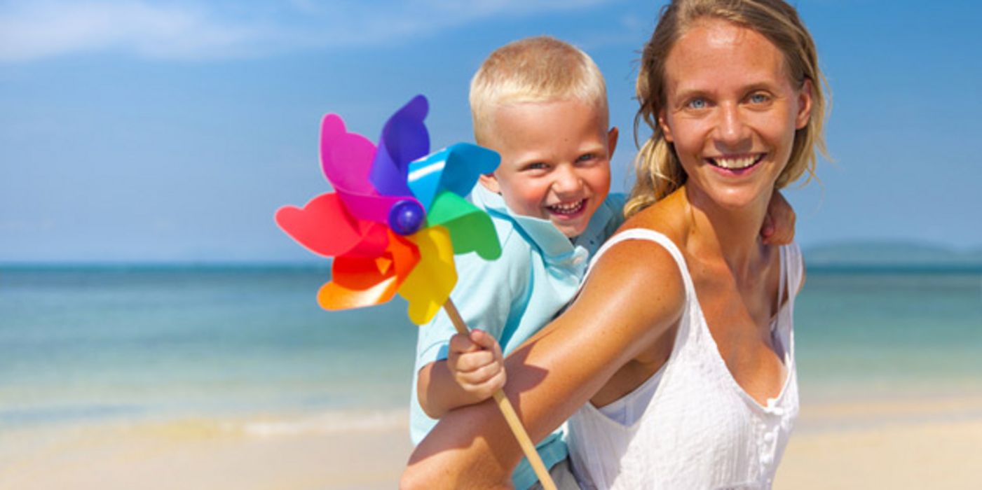 Heiteres Strandfoto: Blonde, lachende Frau, bis Hüfte zu sehen, weißes Shirt, in die Kamera lachend, auf dem Rücken ein kleiner Junge mit blonden Haaren, ein buntes Windrad in der rechten Hand