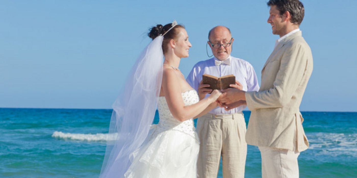 Hochzeitspaar ganz in Weiß sich gegenüberstehend, lächelnd, an den Händen haltend, zwischen ihnen etwas zurückgesetzt Pfarrer (?) auch ganz in Weiß mit rotem Buch, dahinter türkisblaues Meer