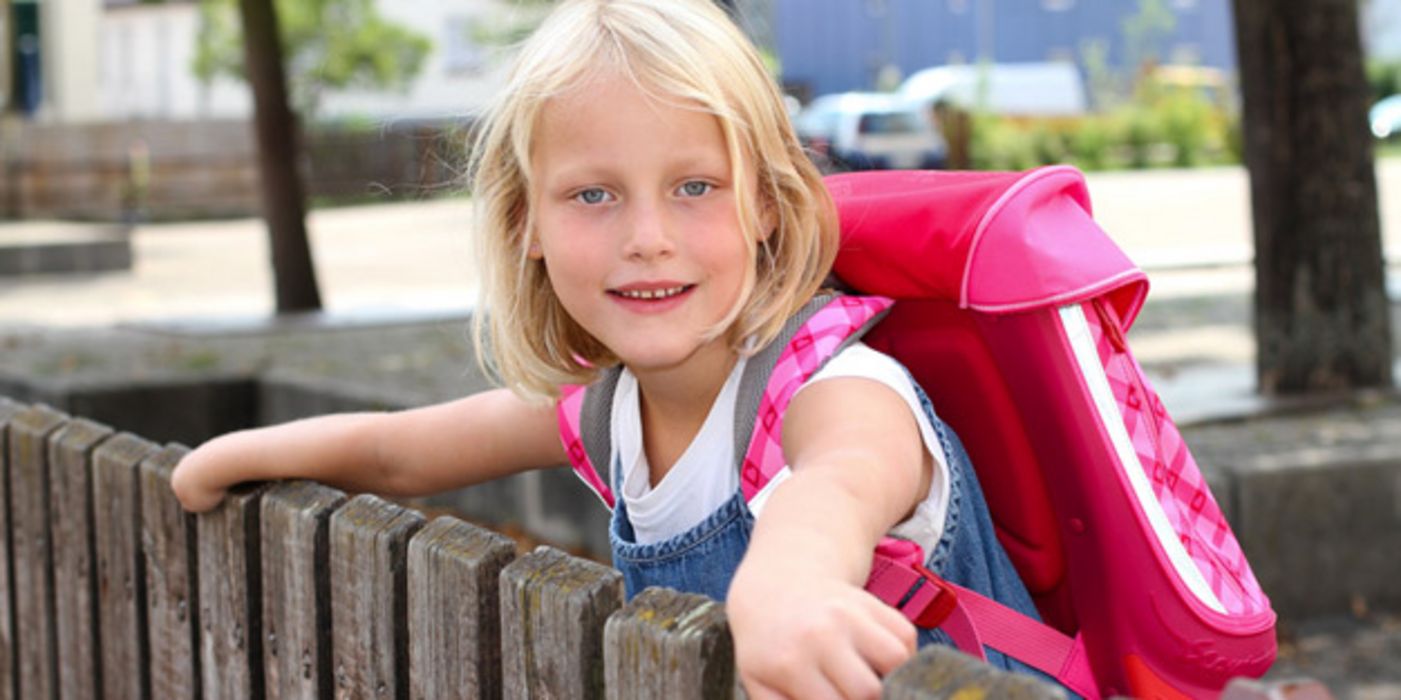 Mädchen, ca. 8 Jahre alt, blond, mit rosafarbenem Schulranzen, über einen Zaun gelehnt und sich dran festhaltend, schaut in die Kamera