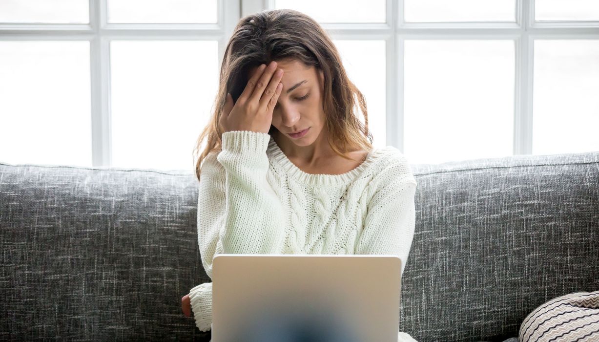 Frau mit Laptop auf dem Schoß, fasst sich an die Schläfe.