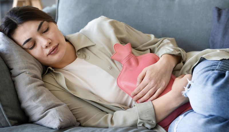 Junge Frau ruht sich auf einem Sofa aus, eine Wärmeflasche auf dem Bauch.