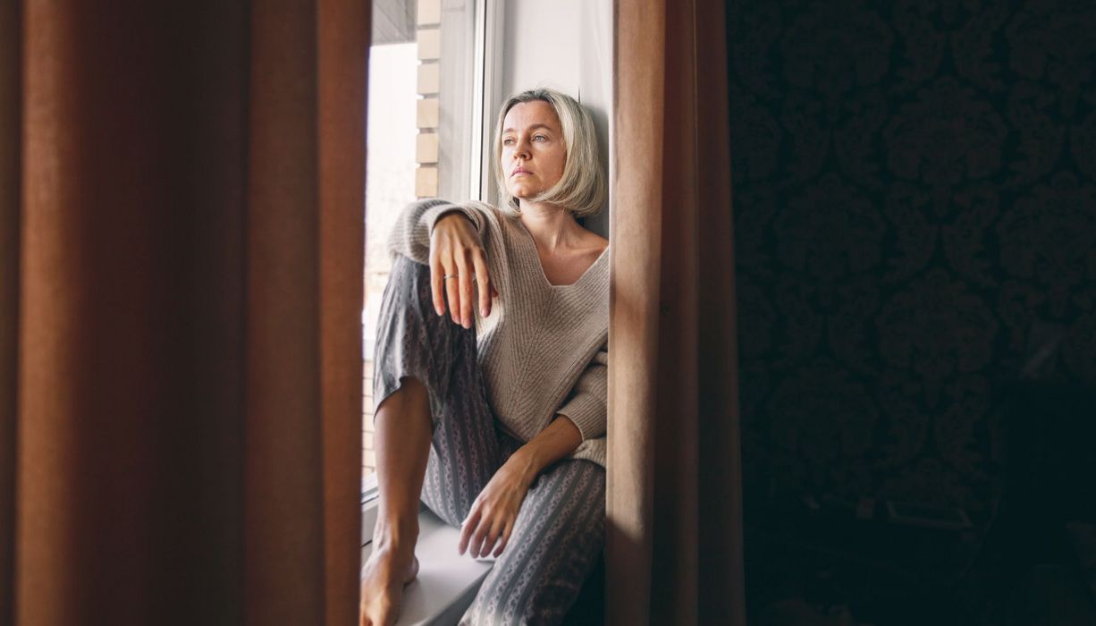 Frau, sitzt auf der Fensterbank und schaut traurig nach draußen.