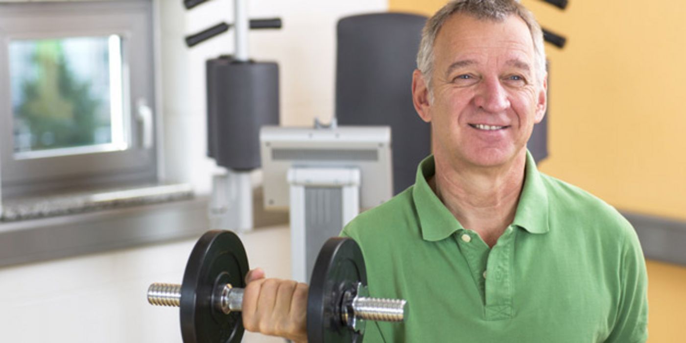 Mann, ca. Mitte 50, grünes Poloshirt, Frontalansicht, mit Hantel in der rechten Hand, gebeugter Arm, Hintergrund Fitnessgeräte im Studio