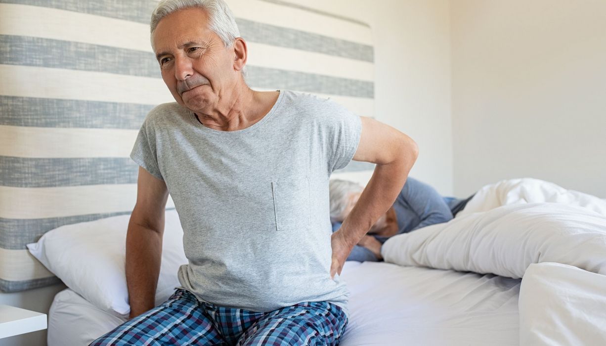 Älterer Mann mit Kreuzschmerzen, sitzt auf seinem Bett.