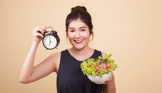 Eine Frau steht vor einem beigen Hintergrund. Sie hält eine Uhr in der rechten Hand und einen Salat in der linken Hand.