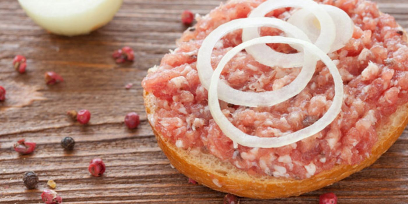 Halbes Mettbrötchen mit Zwiebelringen auf Holztisch, bunte Pfefferkörner, im Hintergrund Zwiebel