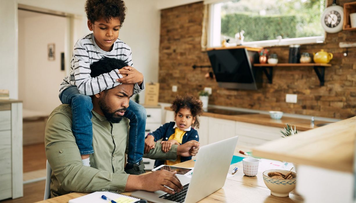 Mann, sitzt im Home Office am Laptop und wird von seinen zwei Kindern gestört.