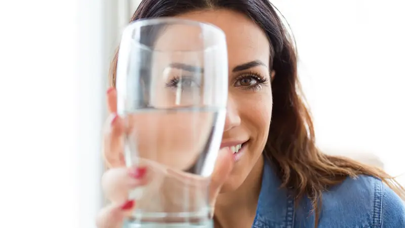 Junge Frau hält ein Glas Wasser dem Betrachter entgegen.