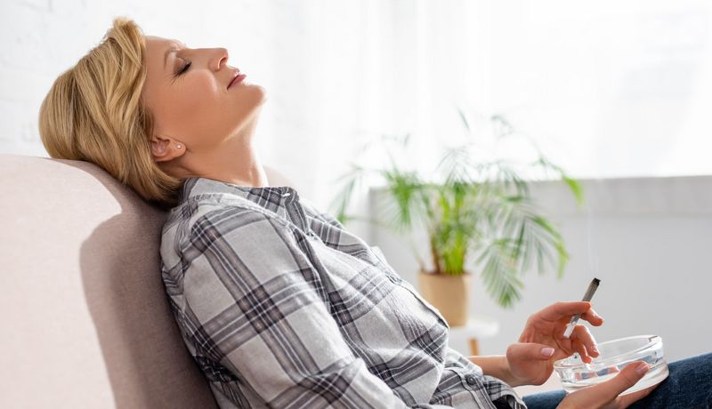 Eine Dame sitzt auf dem Sofa mit nach hinten gelehntem Kopf und hält einen Joint in der Hand.