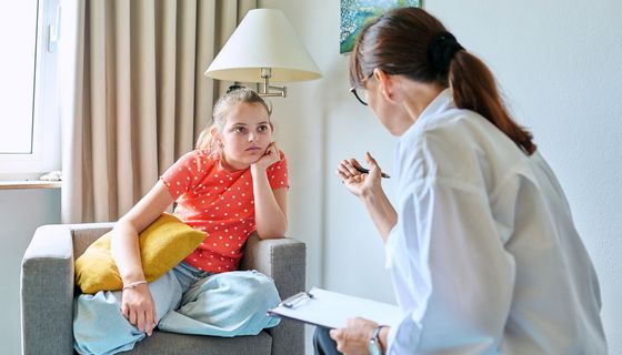 Ein Mädchen, etwa 10 bis 12 Jahre alt, hockt auf einem Sessel und hört einer Therapeutin zu. Sie erklärt ihr etwas, auf dem Schoß ein Klemmbrett.