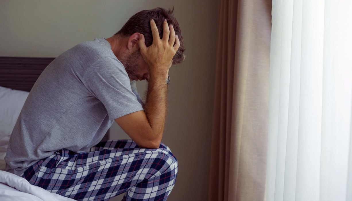 Mann sitzt auf dem Bett, stützt den Kopf in die Hände.