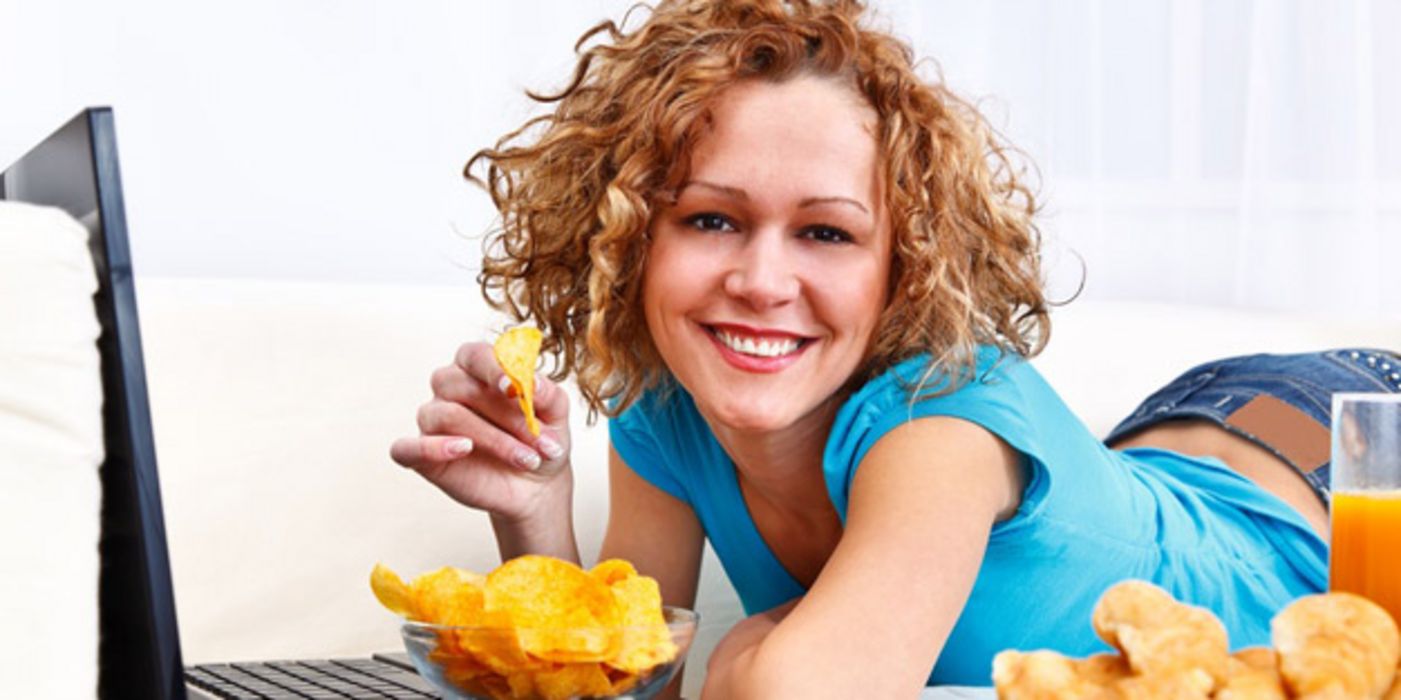 Junge Frau auf dem Bauch liegend vor dem Laptop mit einer Schüssel Kartoffelchips