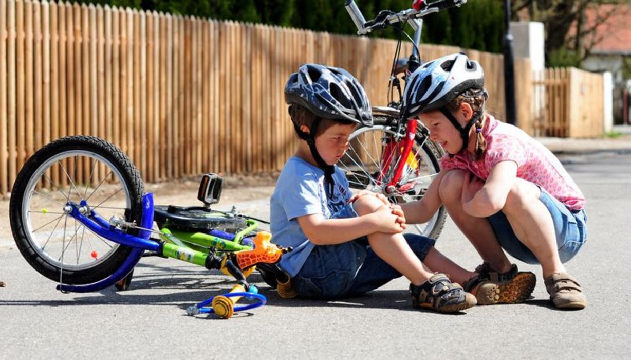 Ein Junge ist vom Fahrrad gefallen und hat sich das Knie aufgeschürft, ein anderer Junge kniet daneben