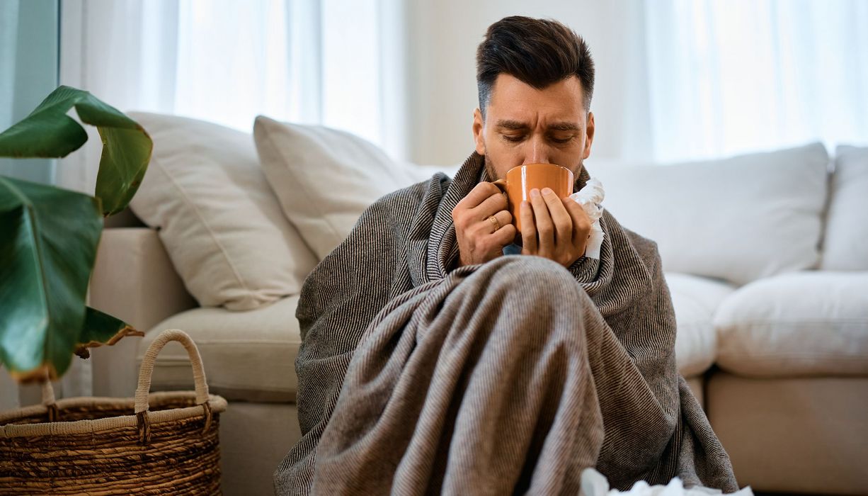 Mann, sitzt mit einer Decke und einer Tasse Tee auf der Couch.