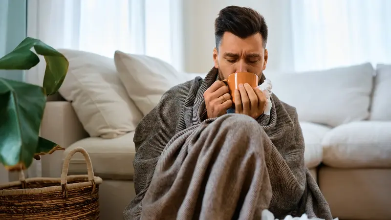 Mann, sitzt mit einer Decke und einer Tasse Tee auf der Couch.