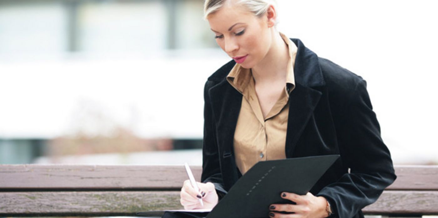 Frau in den 20ern, blonde, zurückgenommene Haare, dunkle Business-Jacke, helle Bluse, auf Bank sitzend in eine Bewerbungsmappe schreibend, ernster Gesichtsausdruck