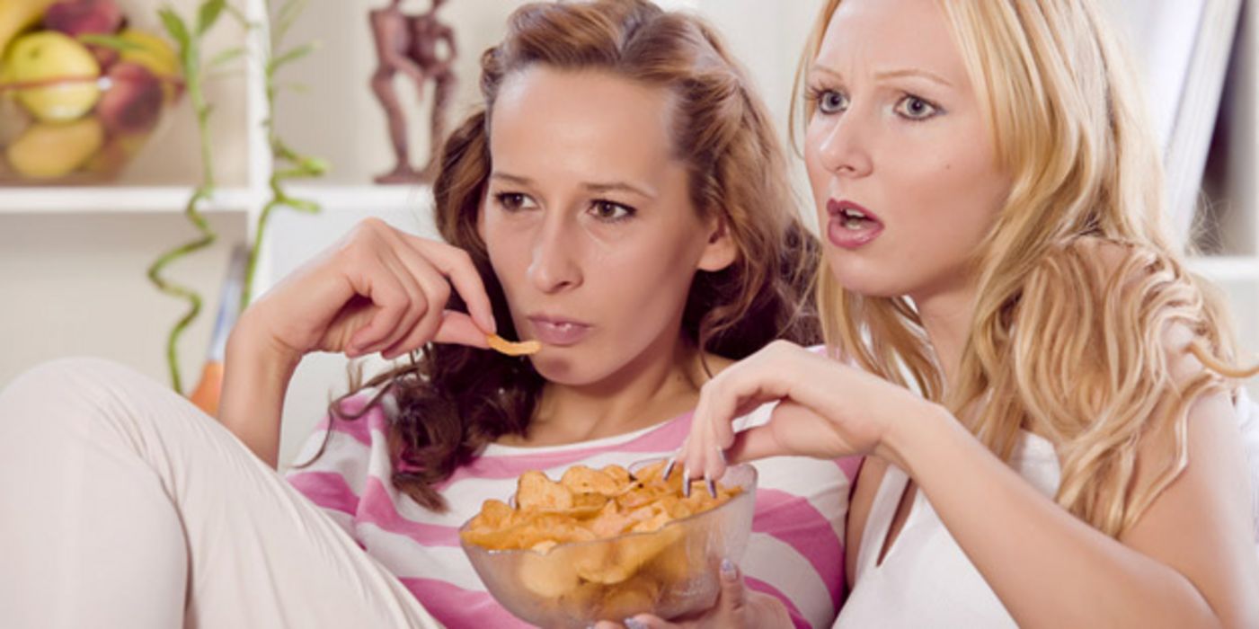 Zwei junge Frauen sitzen zusammen auf der Couch, schauen Fernsehen und essen Chips.