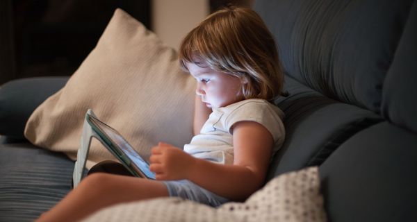 Kleinkind, sitzt mit einem Tablet auf der Couch und schaut auf den Bildschirm.