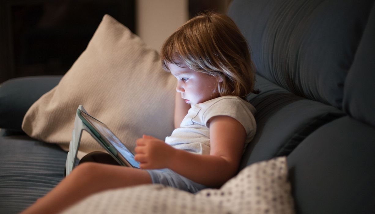 Kleinkind, sitzt mit einem Tablet auf der Couch und schaut auf den Bildschirm.