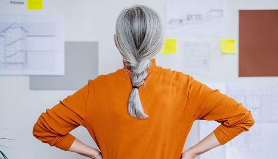 Frau mit grauen Haaren, steht mit dem Gesicht zu einer Wand.