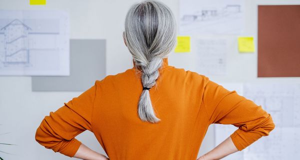 Frau mit grauen Haaren, steht mit dem Gesicht zu einer Wand.
