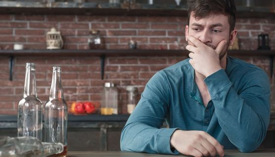 Ein Mann blickt nachdenklich ins Leere, neben ihm stehen zwei fast leere Flaschen, eine liegt umgekippt daneben.