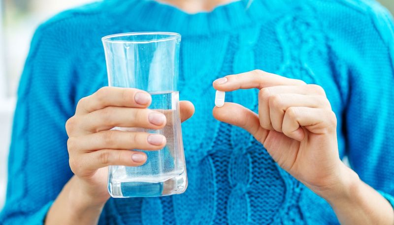 Frau mit blauem Pullover hält ein Glas Wasser und eine Tablette in den Händen.