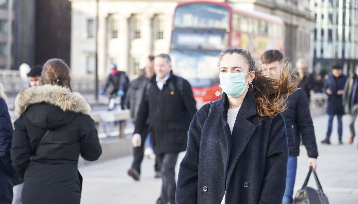 Junge Frau mit Maske, läuft in London auf einem Fußgängerweg, Doppeldecker-Bus im Hintergrund.