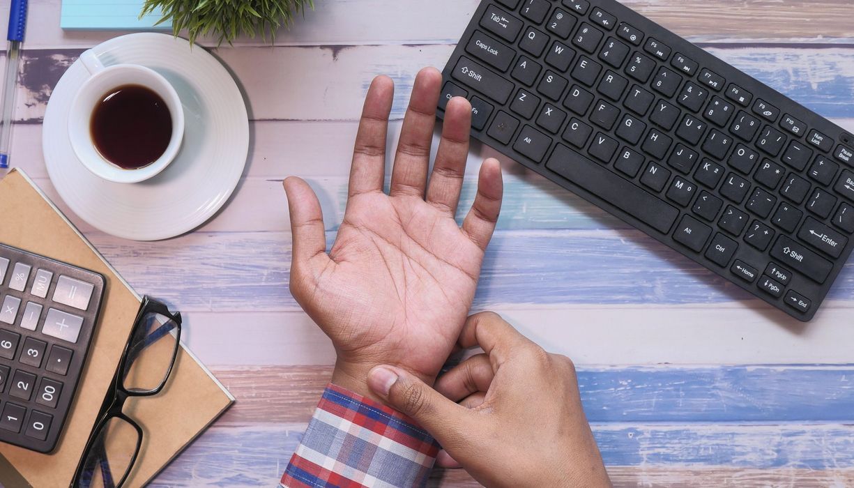 Mann hält sich sein schmerzendes Handgelenk, im Hintergrund eine Tastatur und ein Kaffee.