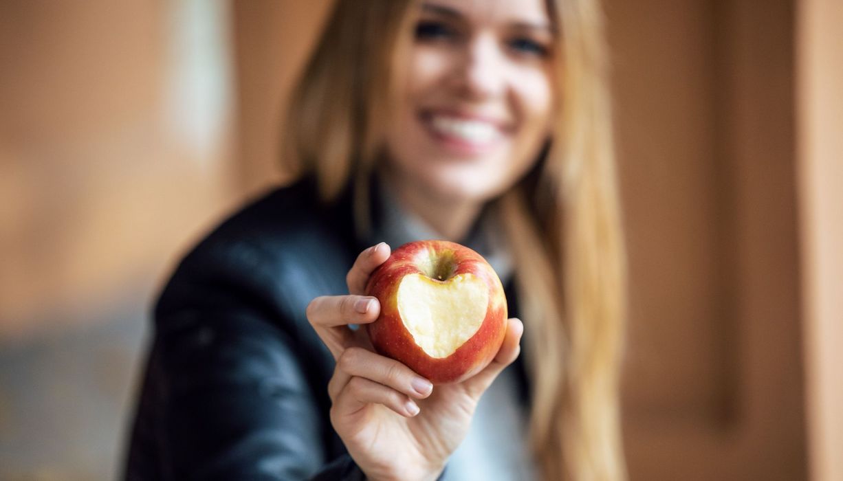 Junge Frau hält einen Apfel in die Kamera, der einen herzförmigen Biss hat.