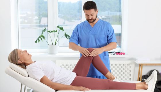 Eine ältere Frau liegt beim Physiotherapeuten auf einer Liege. Ein Bein ist hoch gestützt, der Physiotherapeut hat seine Hände um ihr Knie gelegt.