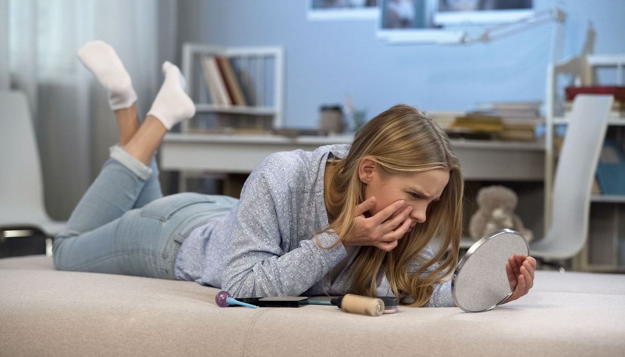 Junges blondes Mädchen, liegt auf dem Boden und betrachtet ihr Gesicht kritisch mit einem Spiegel.