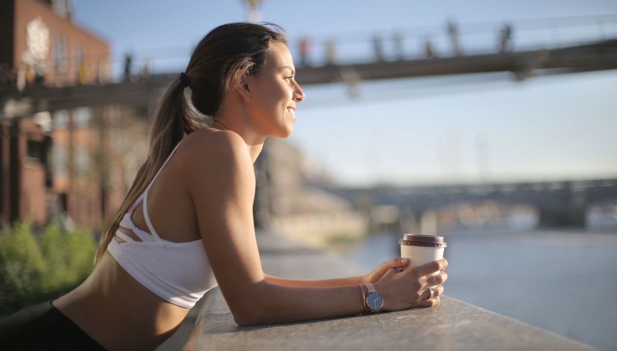 Junge Frau, lehnt sich mit einem Kaffee an eine Brüstung, sportliches Outfit.