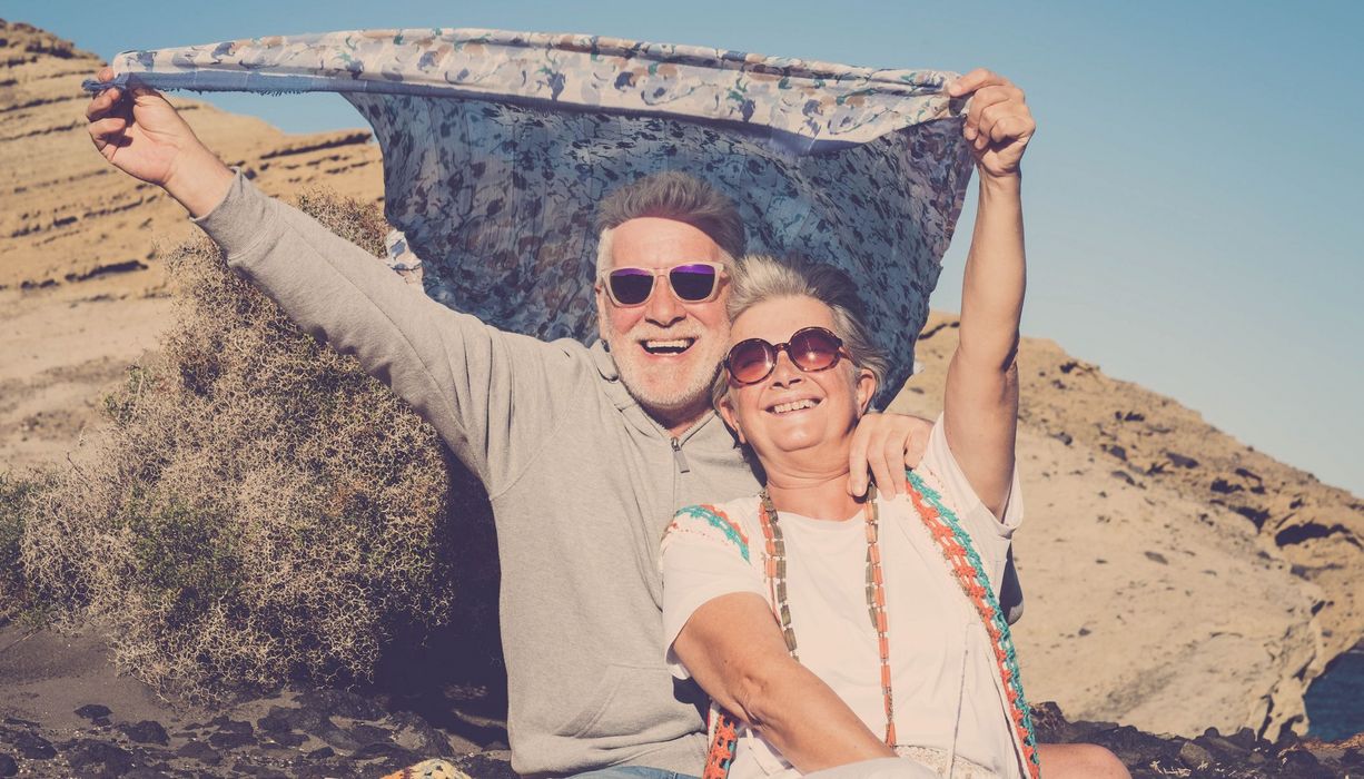 Zwei Senioren am Strand, halten ein Tuch über ihren Kopf und lachen.