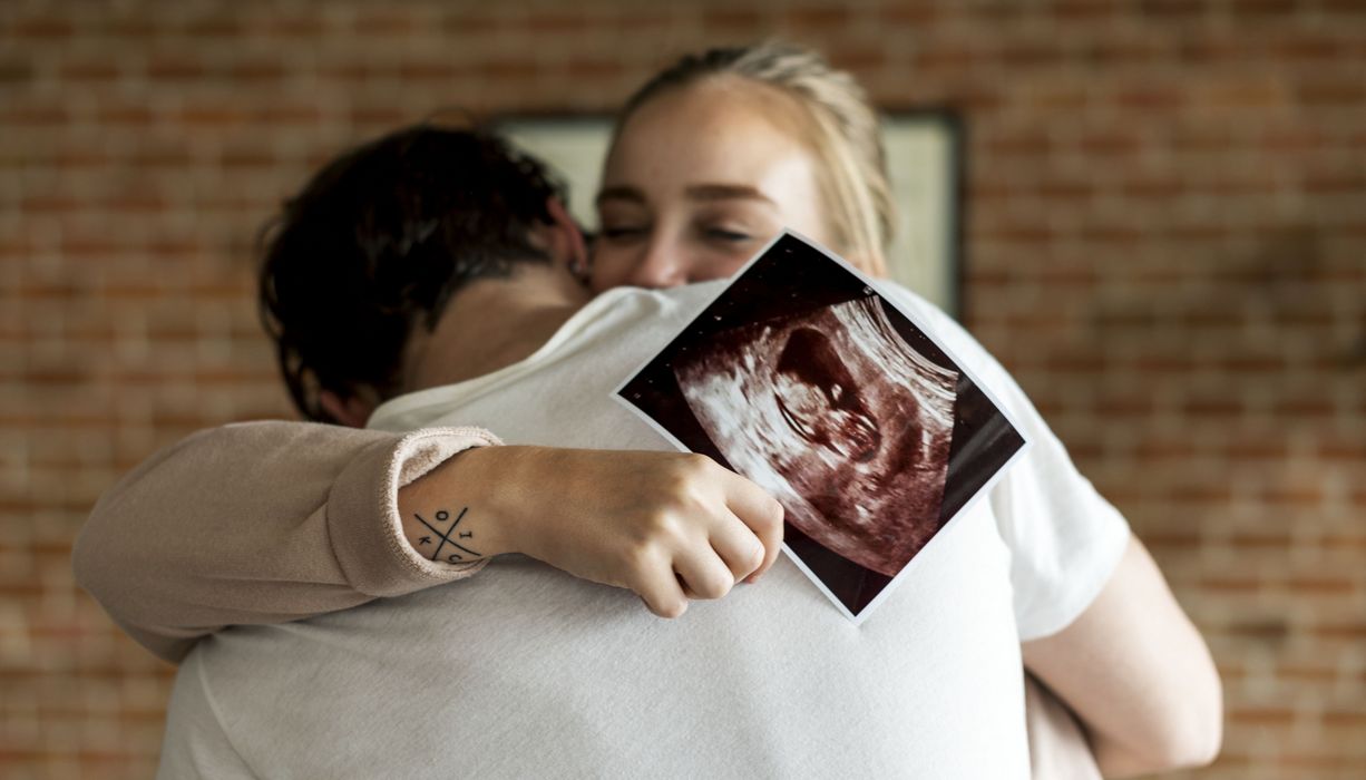 Junge Frau umarmt ihren Partner. In einer Hand hält sie ein Ultraschallbild. 