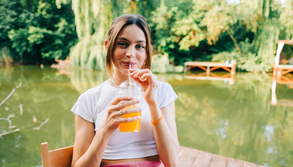 Junge Frau sitzt am See und trinkt Limo.