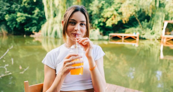 Junge Frau sitzt am See und trinkt Limo.